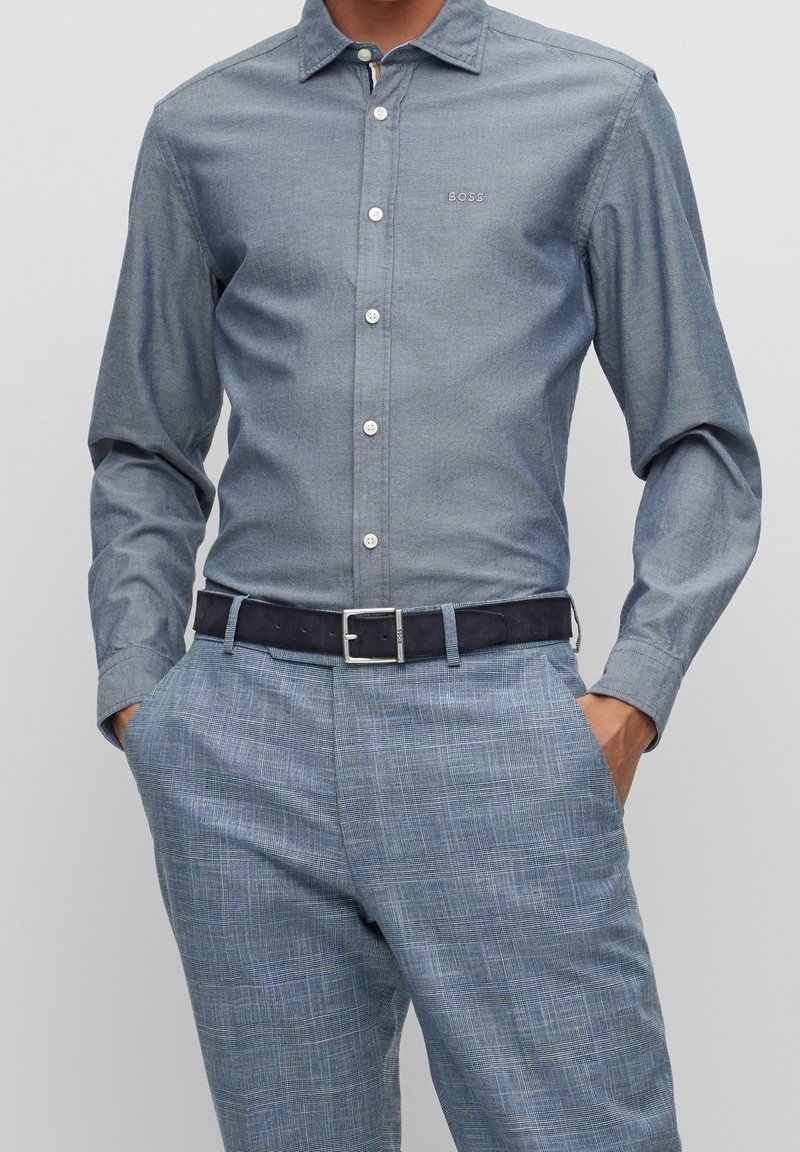 Camicia azzurra chiusa con bottoni, colletto, maniche lunghe e bottoni bianchi, abbinata a pantaloni blu a quadri e cintura scura.