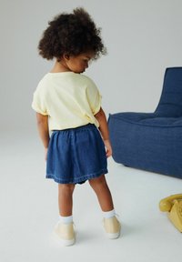 Egy gyermek által viselt sárga rövid ujjú ing és kék farmer szoknya. Fehér bokazokni és világos sárga sportcipő egészíti ki a ruházatot.