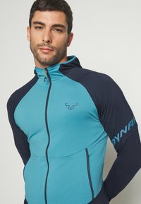 Priliehavá mikina na zips v tyrkysovej a námornej farbe, vyrobená z ľahkej tkaniny. Má malé logo a kontrastné farebné panely na rukávoch.