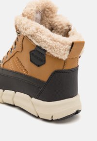 Bruine leren enkellaars met een donzige beige kraag, zwarte rubberen tussenzool en een gestructureerd donker paneel. Heeft een logo-detail en een stevige veterontwerp.