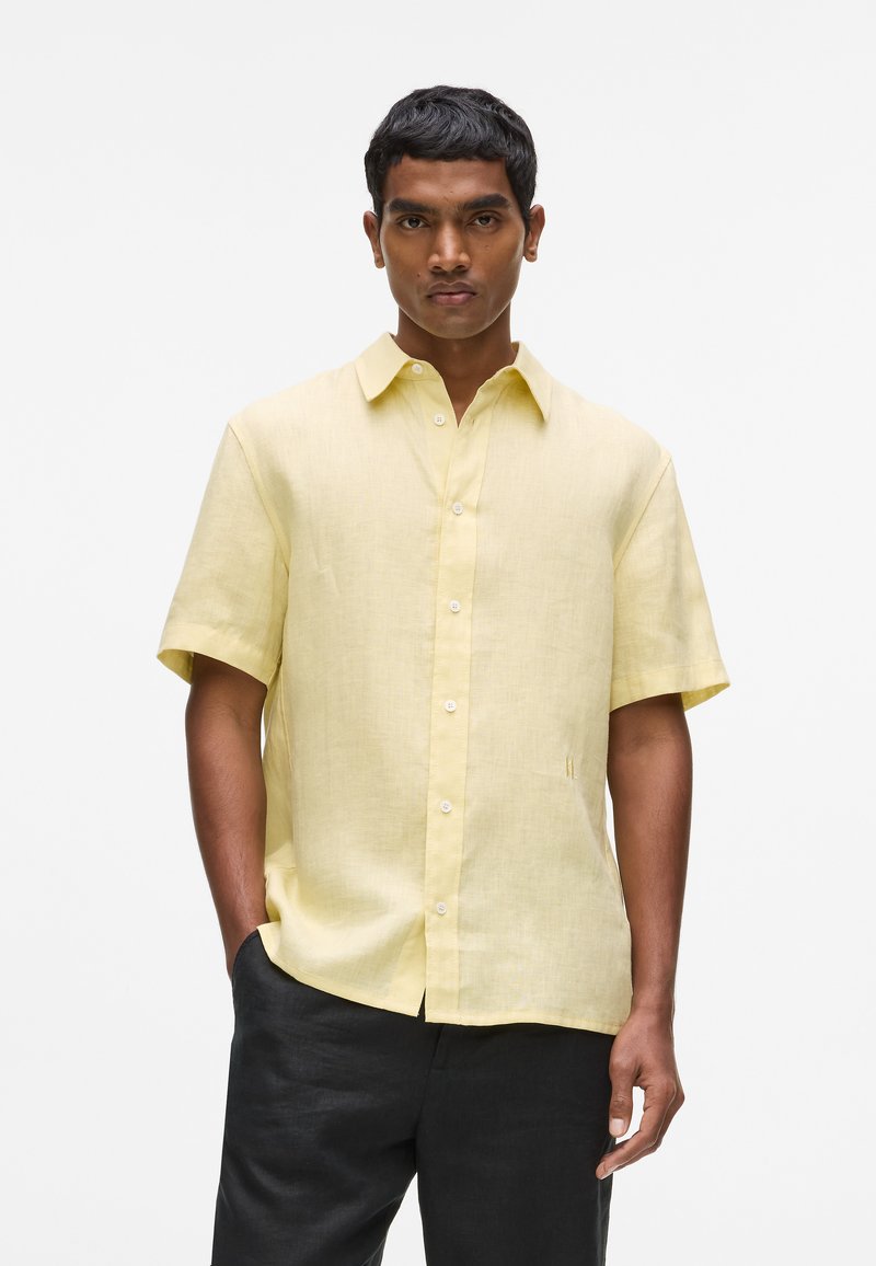 Chemise jaune en lin à manches courtes avec un col boutonné, présentant une texture subtile et un détail de poche latérale, associée à un pantalon foncé.