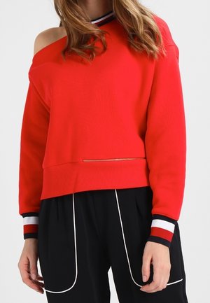 Frau trägt einen roten Sweatshirt mit freiliegenden Schultern, gestreiften Bündchen und schwarze Hose mit weißen Paspeldetails.