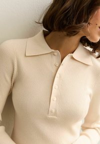 Polo a coste color beige chiaro con colletto morbido e patta con cinque bottoni, caratterizzata da una forma aderente e una texture liscia.