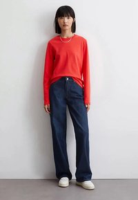 Woman standing against white wall wearing a red long-sleeve shirt, dark blue wide-leg jeans, white sneakers, and a silver necklace.