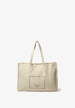 Beige krāsas kanvas soma ar divām plānām rokturiem, kurā ir taisnstūrveida melna grafika ar tekstu "still nordic" iekšpusē.