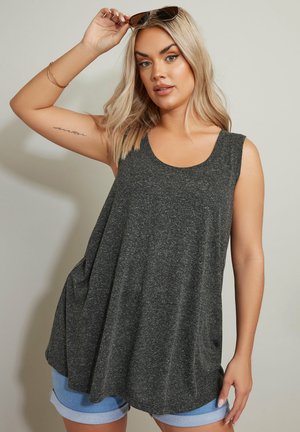 Blondynka w ciemnoszarej, bezrękawnikowej bluzce i podwiniętych szortach z denimu, trzymająca okulary przeciwsłoneczne nad głową, stojąca na jednolitym tle.