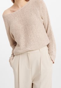 Femme portant un pull en maille beige à épaules dénudées et un pantalon beige taille haute, les mains dans les poches, sur un fond uni.