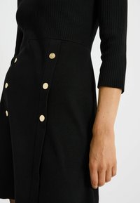 Robe noire en maille avec un corsage côtelé et une jupe évasée, agrémentée de boutons dorés le long de la couture latérale gauche.