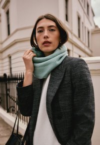 Ribbed, light blue cowl scarf worn over a white sweater and grey blazer. Model stands against a light-colored building with a black fence.