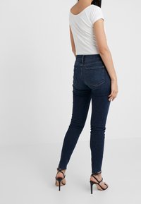 Jeans de mezclilla oscuros de corte slim, con una cintura de altura media, dos bolsillos traseros y una textura suave. Combinados con una camiseta blanca básica y sandalias negras de tacón.