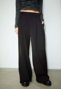 Zwarte broek met wijde pijpen en een zachte textuur, voorzien van een knoopsluiting, voorplooien en zakken. Draagt een gestreepte top met lange mouwen.