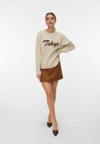 Beige sweater met "Tokyo" in bruine letters, gecombineerd met een bruine minirok en puntige hakken met decoratieve studs.
