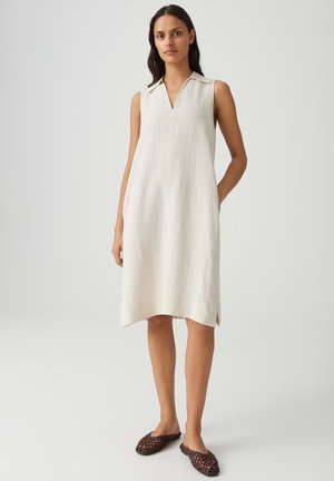 Donna in piedi che indossa un vestito senza maniche, lungo fino al ginocchio, color avorio con colletto e scarpe marroni intrecciate senza lacci, su sfondo neutro.