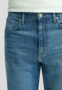 Primo piano di jeans in denim blu con tasche frontali, passanti per cintura e vita con bottone indossati con una camicia bianca.