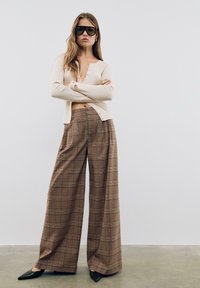 Cardigan beige à côtes avec détails de boutons associé à un pantalon large en vichy marron et des escarpins noirs à bout pointu. Modèle portant des lunettes de soleil oversize.
