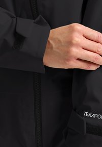 Manche de veste imperméable noire avec fermeture Velcro. La marque visible indique "TEXAPORE". Présente une texture lisse et un design élégant.