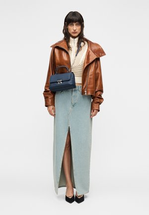 Γυναίκα που φορεί ένα καφέ δερμάτινο μπουφάν, κρεμ πλεκτό πουλόβερ, μπλε τζιν φούστα με σκίσιμο μπροστά, μαύρες γόβες και κρατά μια μπλε σκούρη τσάντα ώμου στο στήθος της.