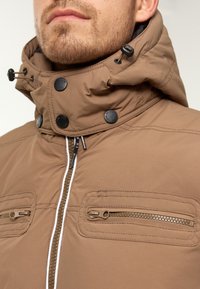 Mann trägt eine braune Jacke mit hohem Kragen, verstellbarem Kapuze, Frontrei�ßverschluss und zwei horizontalen Reißverschlusstaschen auf der Brust.
