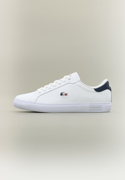 Бели маратонки Lacoste с нисък профил, с тъмен син хавлиен етикет на задната част, бели връзки, брандиран крокодилски логотип отстрани и бяла гумена подметка.