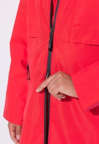 Czerwona wodoodporna kurtka z czarnym zamkiem błyskawicznym, wyposażona w przedni zamek oraz kieszenie po bokach. Gładka tekstura i luźny krój.