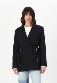 Črn blazer s prilagojeno zasnovo, ki ima dva sprednja žepa, prilagodljive vrvice in gladko teksturo tkanine.