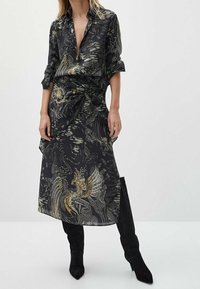 Black shirt dress with a deep v-neck, knotted waist, and intricate gold and white patterns. Paired with knee-high black boots.