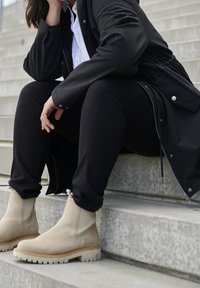 Beige Wildleder-Stiefeletten mit dicken weißen Sohlen, kombiniert mit schwarzen engen Hosen und einer schwarzen Jacke, sitzend auf Betontreppen.