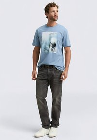 Šviesiai mėlynas medvilninis marškinėlis su grafiniu atspaudu centre priekyje, derinamas su tamsiomis džinsinėmis kelnėmis ir baltais sportbačiais. Trumpomis rankovėmis, apvalia iškirpte.