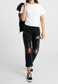 T-shirt bianco corto, jeans neri strappati con orlo frangiato e buchi sulle ginocchia, e sneakers Vans nere, abbinati a una giacca nera.