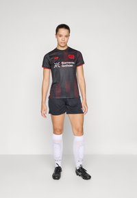Čierno-červený pruhovaný futbalový dres s logom, skombinovaný s čiernymi šortkami. Biele ponožky po kolená a čierne kopačky dopĺňajú vzhľad.