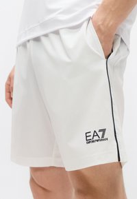 Biele športové šortky s bočnými vreckami, čiernym lemovaním a logom EA7. Vyrobené z ľahkého materiálu s hladkou textúrou.