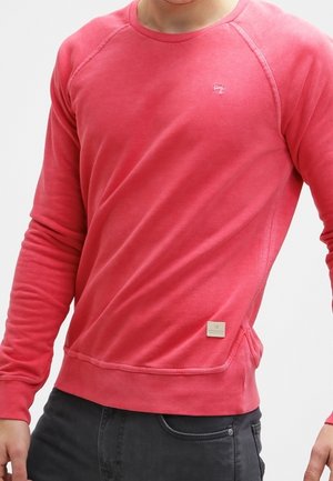 Rode lange mouwen trui gedragen door een persoon, gecombineerd met donkergrijze broek, met een klein logo op de borst en een stoffen label bij de zoom.