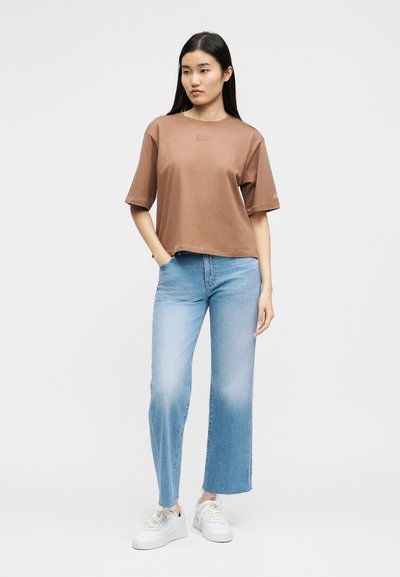 Νεαρή γυναίκα στέκεται με το χέρι στην τσέπη, φορώντας ένα φαρδύ καφέ t-shirt, ανοιχτού μπλε τζιν και λευκά αθλητικά παπούτσια, πάνω σε απλό φόντο.