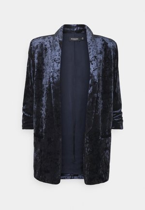 Sammalepukki tummanlaivastonsinisestä sametista, jossa on pehmeä pinta, avoin etuosa, kolmen neljäsosan hihat ja kaksi sivutaskua lisäyksityiskohtana.