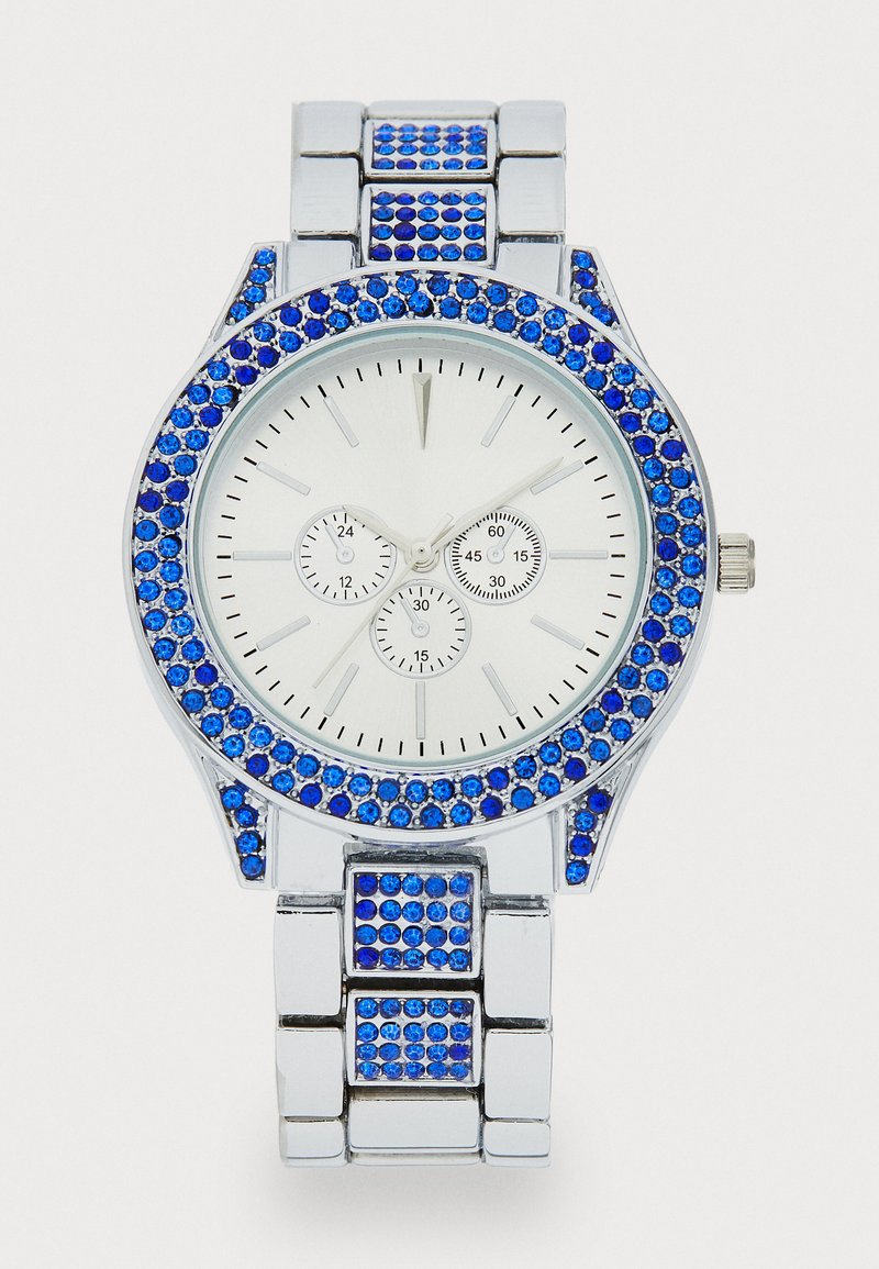 Reloj plateado con acentos de cristal azul alrededor del bisel y la correa. Presenta tres diales más pequeños en una esfera blanca. Diseño metálico y audaz.