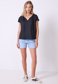 Blusa nera a maniche corte con scollatura a V e dettaglio con cordoncino, abbinata a pantaloni corti in denim blu chiaro e scarpe da ginnastica bianche con accenti neri.