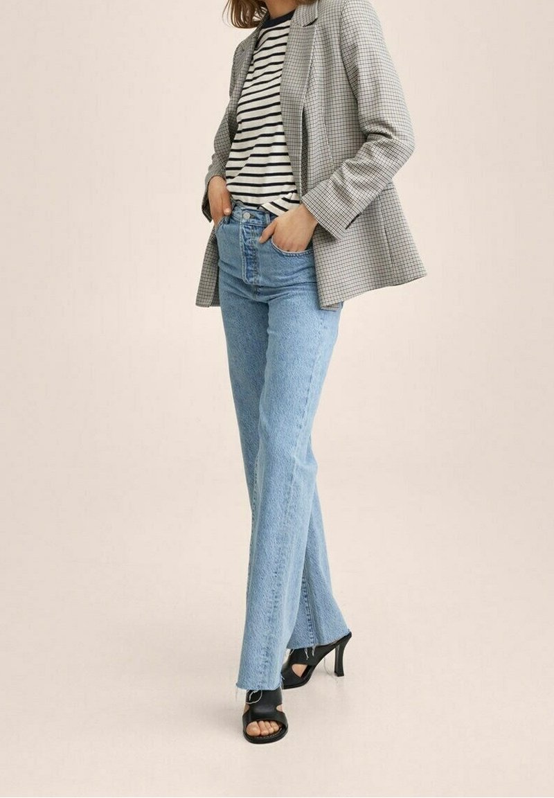 Femme portant un jean taille haute bleu clair, des talons noirs ouverts, une chemise rayée et un blazer à carreaux, les mains dans les poches, sur fond uni.
