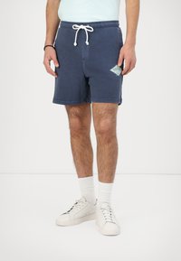 Marineblå bomuldsshorts med snor i taljen, sidelommer, logo-patch og en afslappet pasform. Båret med hvide sneakers og sokker.