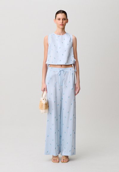 Top corto azzurro chiaro e pantaloni a gamba larga abbinati con piccoli motivi floreali. La modella tiene una piccola borsa intrecciata e indossa sandali chiari.