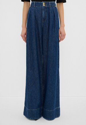 Jean en denim bleu foncé taille haute à jambes larges, avec plis sur le devant et fermeture double bouton, porté avec un haut noir.