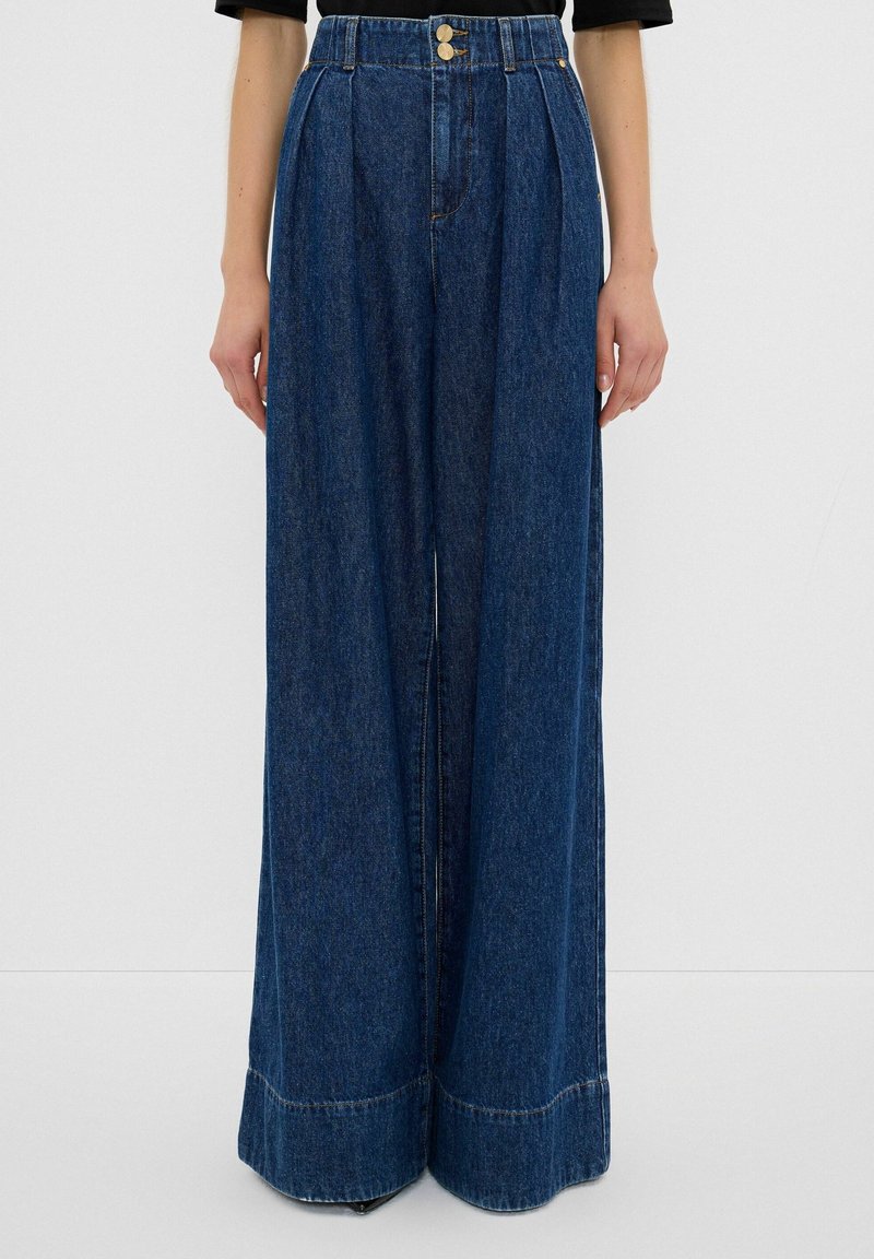 Jean en denim bleu foncé taille haute à jambes larges, avec plis sur le devant et fermeture double bouton, porté avec un haut noir.