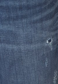 Tela de denim azul con una textura desgastada y ligero desgaste. Presenta un pequeño desgarro y costuras irregulares, manteniendo un aspecto casual.