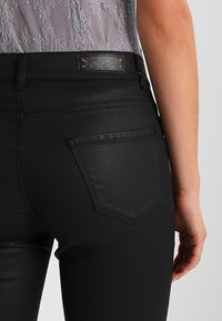 Jeans noirs avec une texture lisse et mate. Dotés d'une étiquette en cuir au niveau de la taille et d'une seule poche arrière avec un motif de coutures en losange.