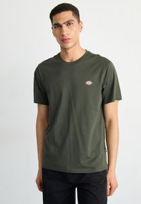 Tricou din bumbac verde închis, cu guler rotund, mâneci scurte și un mic logo pe pieptul stâng, având o margine dreaptă și o textură moale.