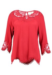 Blusa rossa con maniche a tre quarti, dettagli in pizzo sul colletto e sui polsini, orlo ondulato e rifinitura con pompon lungo il bordo inferiore.