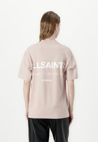 Rosa bomull T-skjorte med korte ermer, med hvit tekst på baksiden: "ALLSAINTS," japanske tegn, og "LONDON." Glatt tekstur.