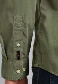 Camicetta verde chiaro con polsini a bottoni, dettagli cuciti e una piccola etichetta del marchio vicino al polsino; il tessuto appare liscio e leggero.
