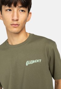 Olivgrünes Baumwoll-T-Shirt mit kurzen Ärmeln und Rundhalsausschnitt, mit einem hellblauen "ELEMENT"-Logo, das auf der Vorderseite gedruckt ist.