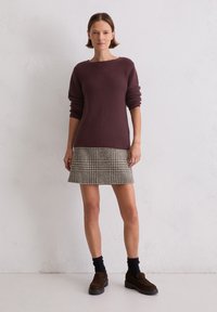 Bordowy sweter z dzianiny, spódnica w wzór houndstooth w odcieniach beżu i szarości, czarne skarpetki i brązowe buty typu slip-on na tle teksturowanej białej ściany.