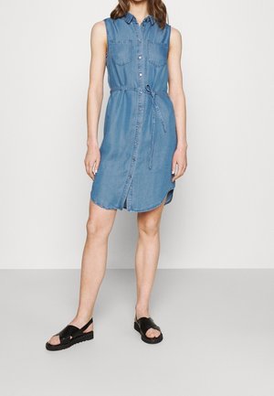 Niebieska sukienka koszulowa bez rękawów z denimu, z przodu zapinana na guziki, z kieszeniami na piersi i wiązaniem w talii, noszona z czarnymi sandałami z odkrytymi palcami na jednolitym tle.